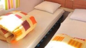 Un llit individual amb llençols estriats, un panell de fusta al capçal i una finestra amb persianes verticals.