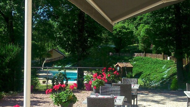 An outdoor patio with wicker furniture, a covered area, and a view of a pool and greenery.