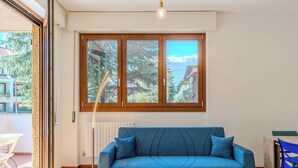 Un salon avec un canapé bleu, une table avec des tasses, une fenêtre donnant sur des arbres et des immeubles, et un radiateur.