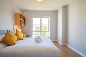 A bedroom with a bed, two pillows, a bedside table, a lamp, a shelf with a plant, and a window. - Large Townhouse with Parking by Shortstays (Galway)