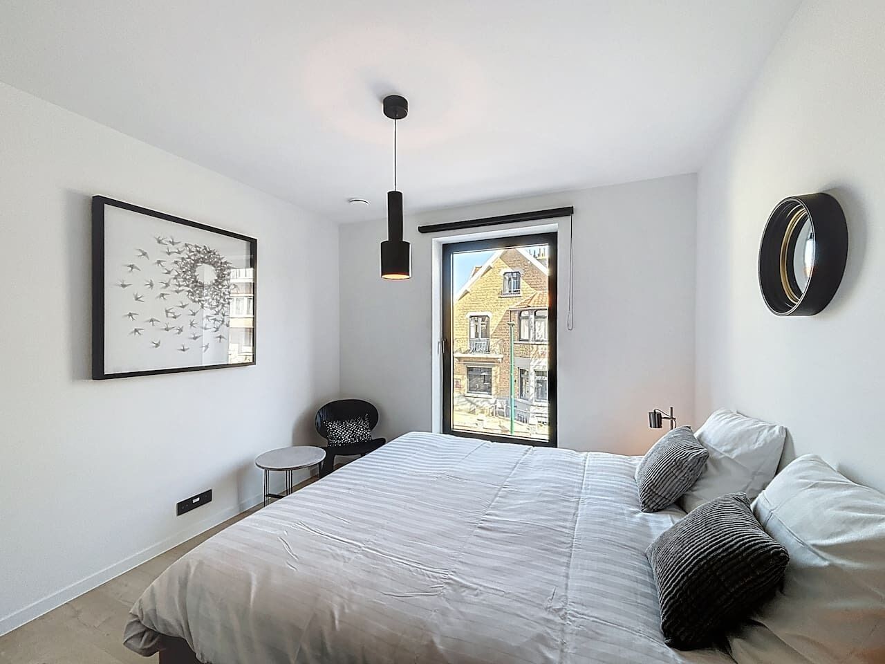 Een moderne slaapkamer met een groot bed, een klein rond tafeltje, een stoel, een kunstwerk in een lijst, een hanglamp en uitzicht op gebouwen door een raam.