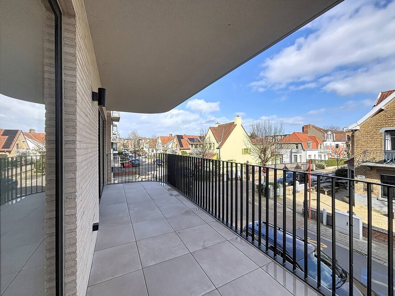Een balkon met uitzicht op een woonstraat, een metalen hekwerk en een heldere lucht.