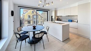 A modern kitchen with a large island, a dining area with a round table and chairs, and a view of buildings through large windows. - Flat Seadune - De Panne (De Panne)