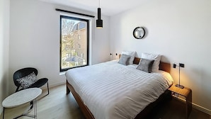 A bedroom with a large bed, a chair, a bedside table, a lamp, and a window with a view of buildings. - Flat Seadune - De Panne (De Panne)