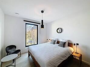 Une chambre à coucher avec un grand lit, une chaise, une table de chevet, une lampe et une fenêtre donnant sur des immeubles.