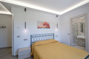 A bedroom with a bed, bedside tables, a wall-mounted painting, and a bathroom with a patterned shower door. - Villavità rooms&more (Pianillo)