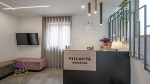 Een receptiebalie in een hotel met een klok, een flatscreen televisie, een bank en een raam met gordijnen.