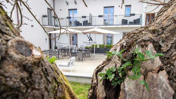 Een gebouw met een bord waarop 'VILLAVITA rooms & more' staat, buiten zitplaatsen met parasols en een boom op de voorgrond.