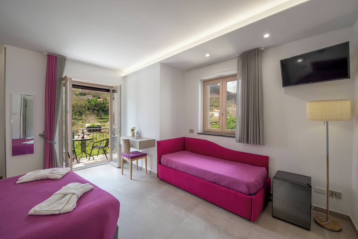 Habitació d'hotel amb un llit, un sofà rosa, un escriptori amb una cadira, una televisió i un balcó amb taula i cadires.