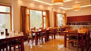 A dining area with multiple wooden tables and chairs, large windows with curtains, and a kitchen area in the background. - Hotel Walnut Ladakh (Leh)