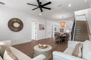 A spacious living room with a high ceiling fan, a dining area with wooden furniture, and a staircase leading to another level. - New Destin Beach Home | Pool | Walk to Beach! (Destin)