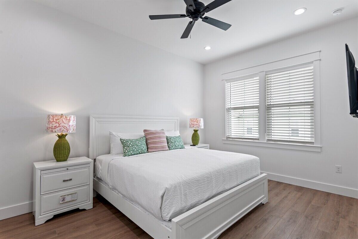 A bedroom with a white bed, two nightstands, a ceiling fan, and a window with blinds.