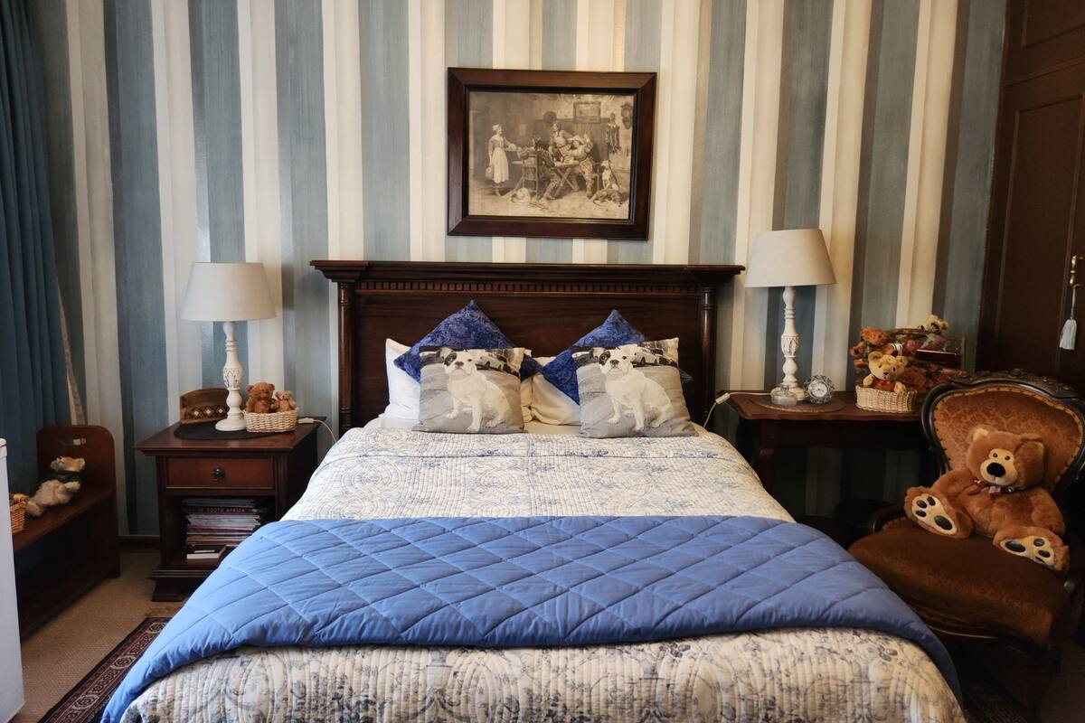 A bedroom with a striped wallpaper, a wooden bed frame, two bedside lamps, a framed picture on the wall, and a chair with teddy bears.