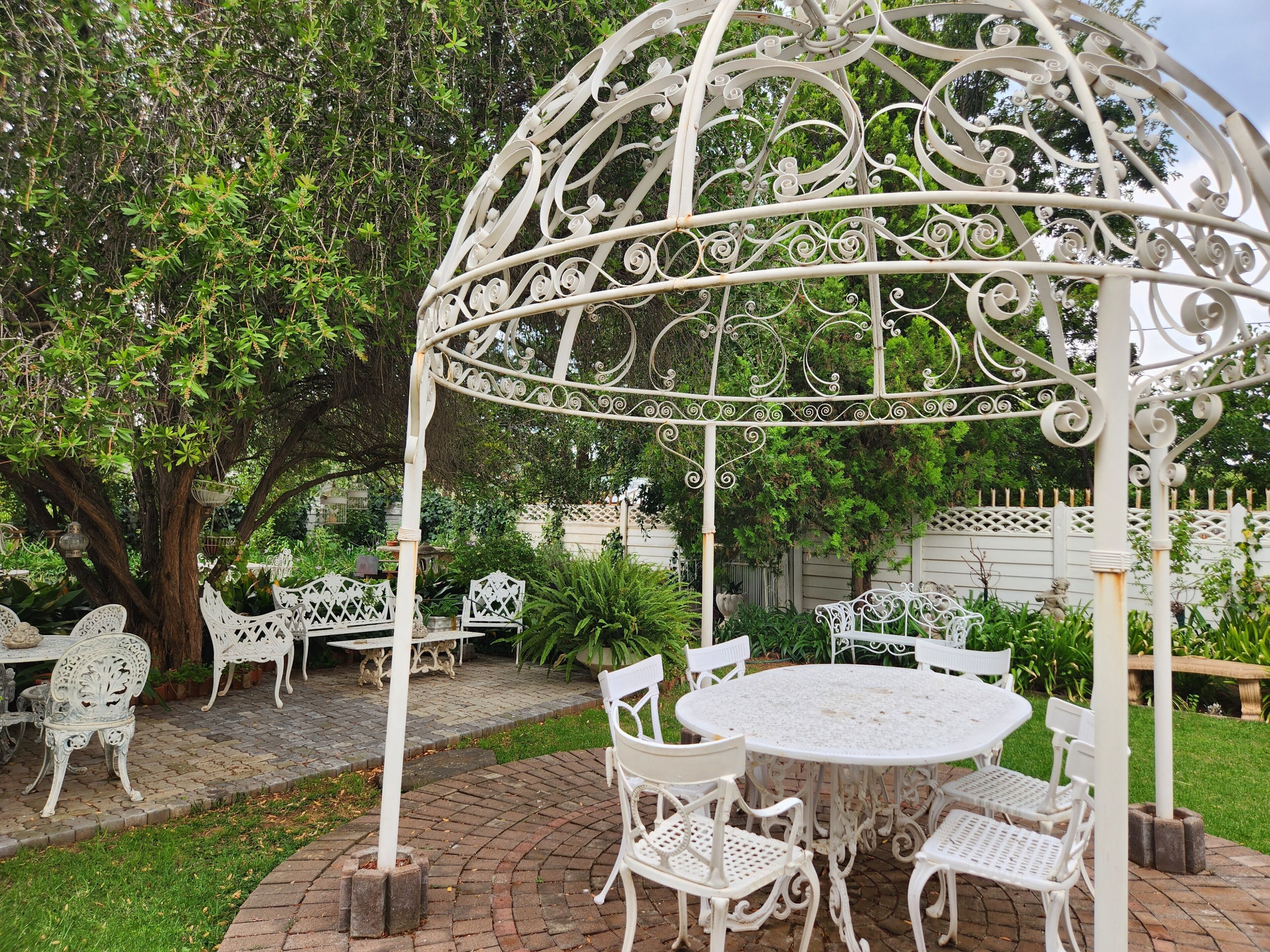 Un jardin agrémenté d’un belvédère en métal blanc, de chaises blanches et d’une table ronde.