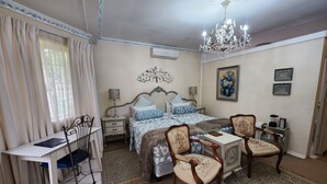 A bedroom with a bed, two chairs, a desk, a chandelier, and a painting on the wall. - THE SUMMER PLACE BOUTIQUE GUESTHOUSE (Bloemfontein)
