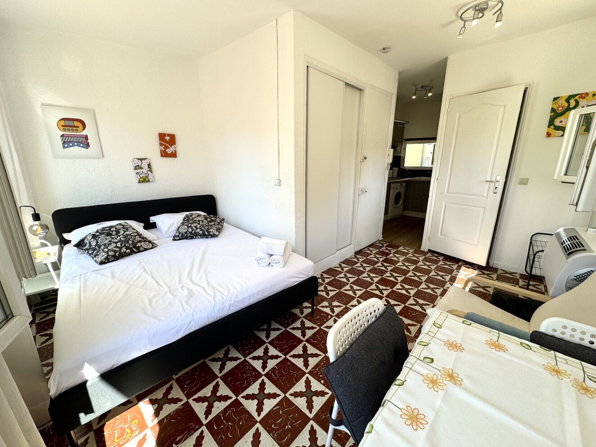 A bedroom with a bed, a chair, a table, and a kitchen area in the background.