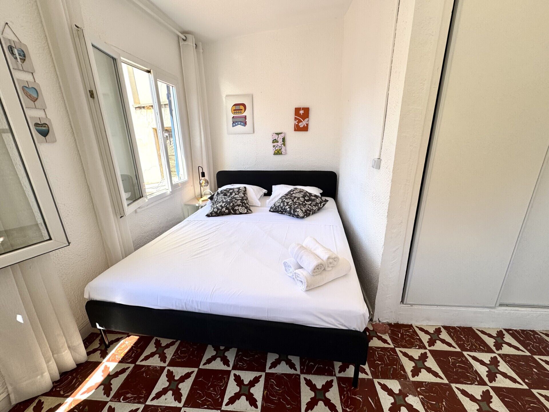 Un lit bien fait, avec du linge de lit blanc et deux coussins à motifs, une table de chevet avec une lampe, et un motif à carreaux sur le sol.