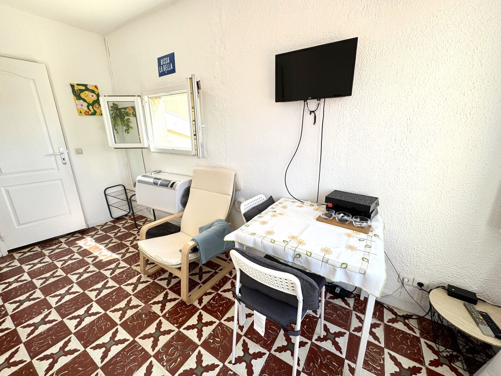 A room with a tiled floor, a chair, a table with a floral tablecloth, a wall-mounted TV, and a heater.
