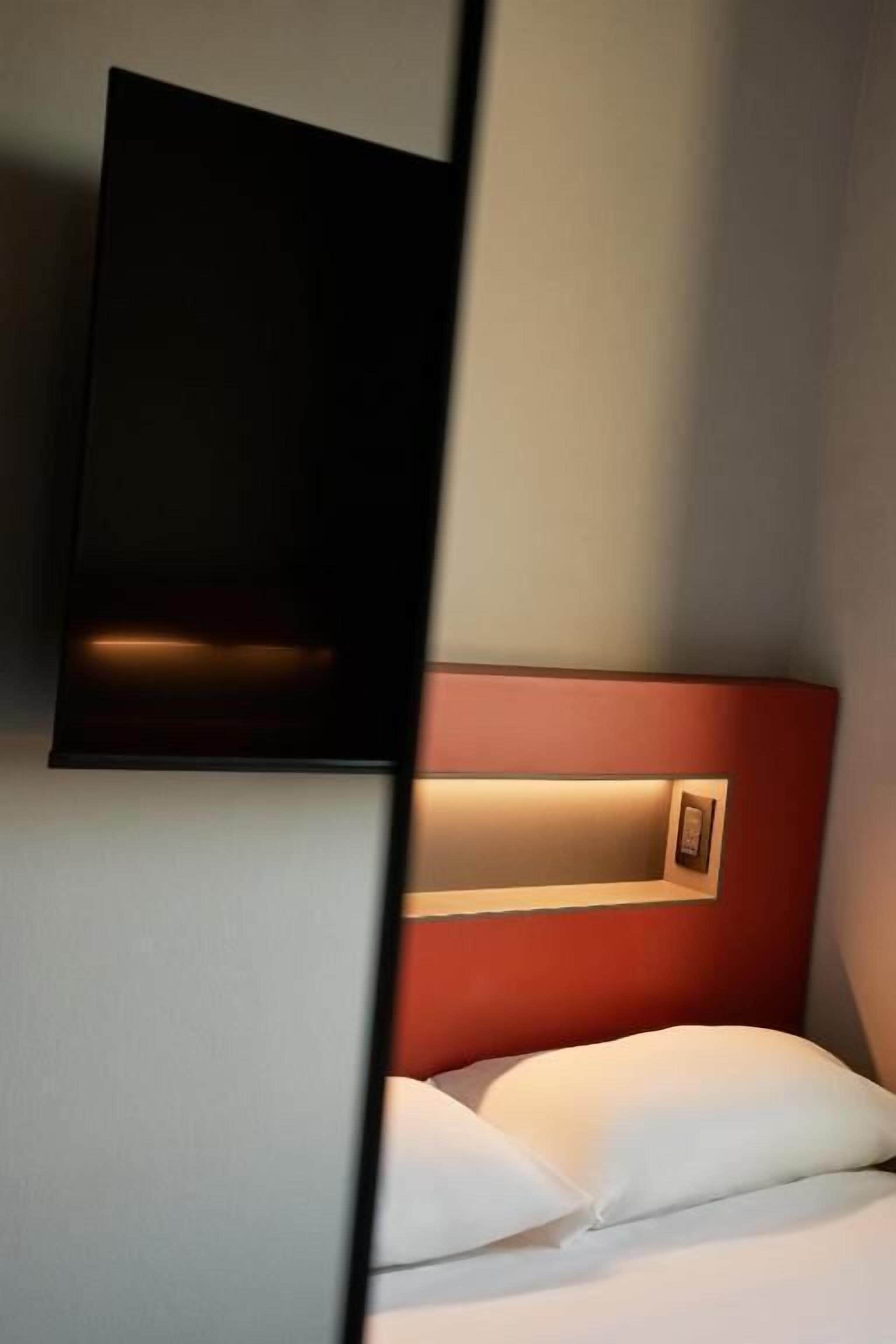 A hotel room with a bed, a wall-mounted TV, and a bedside table with a lamp.