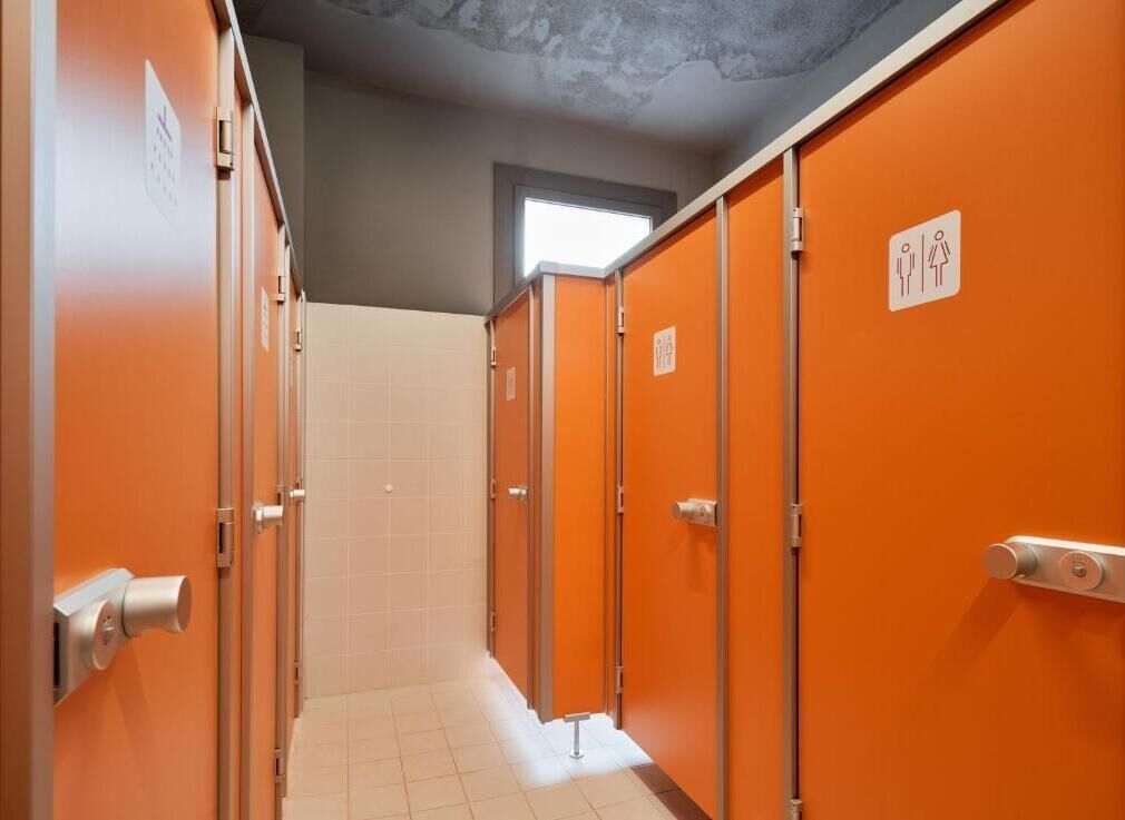 Une rangée de toilettes publiques aux portes orange, avec un sol carrelé et une fenêtre au-dessus.