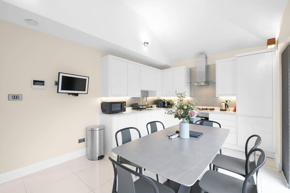 A modern kitchen with white cabinets, a dining table with chairs, a TV mounted on the wall, and a trash bin. - Modern Comfort 2-Bedroom, 2-Bathroom House (Chessington)