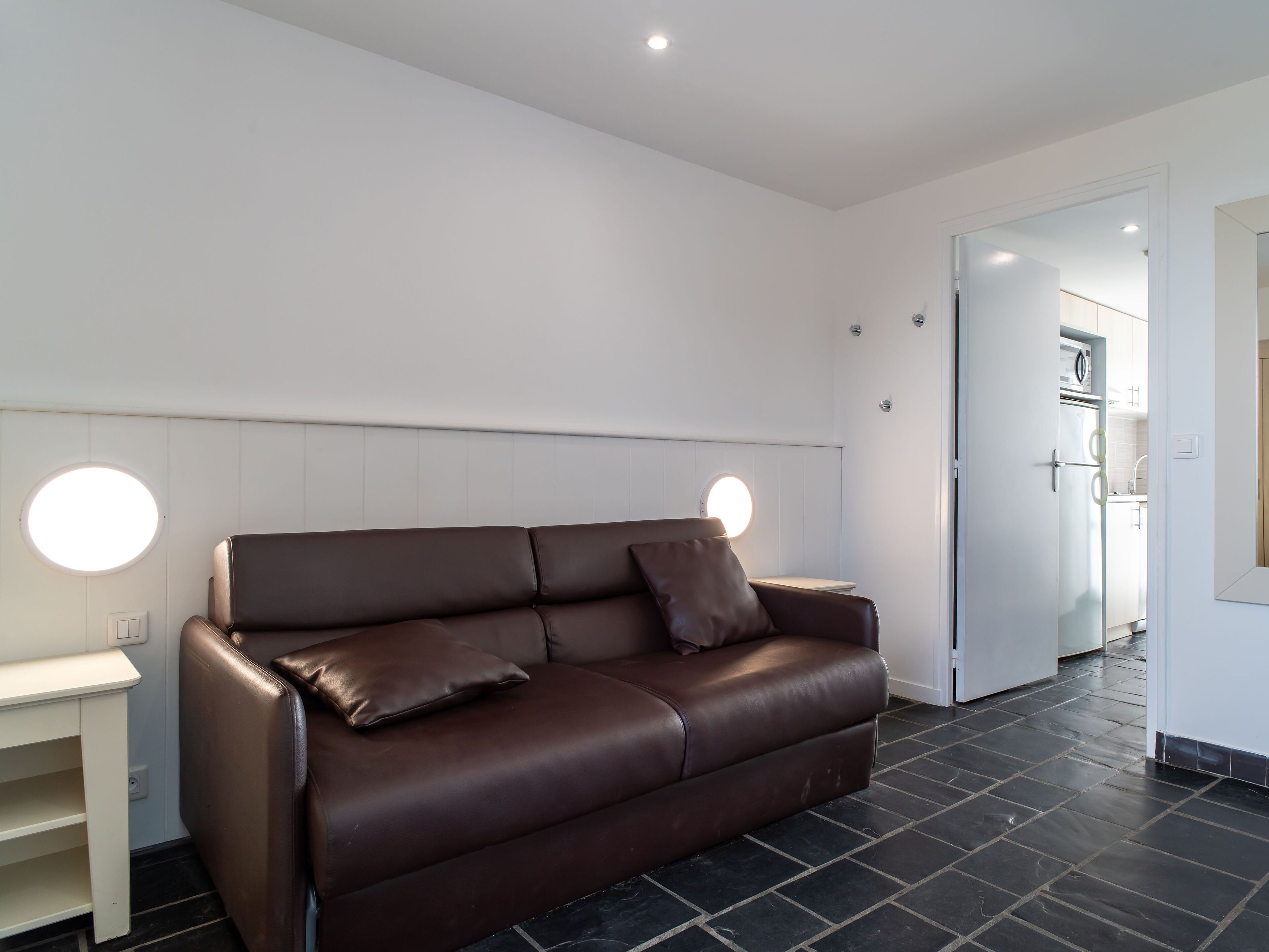 A modern living room with a brown leather sofa, two round wall sconces, and a kitchen visible through an open door.