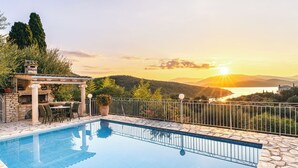 En pool med stentile, en pergola med siddepladser og udsigt til bakker og en solnedgang.