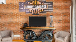 Une pièce avec un mur en briques, une enseigne Harley-Davidson, un téléviseur, une lampe et deux chaises.