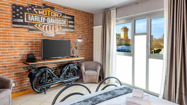 Une chambre d’hôtel avec un mur en briques, une enseigne Harley-Davidson, une télévision, un vélo, une chaise, un lit et une vue sur un château par la fenêtre.
