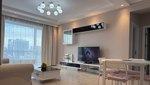Ein modernes Wohnzimmer mit Flachbildfernseher, Glastisch, Sofa, Esstisch mit Stühlen und Stadtblick durch große Fenster.