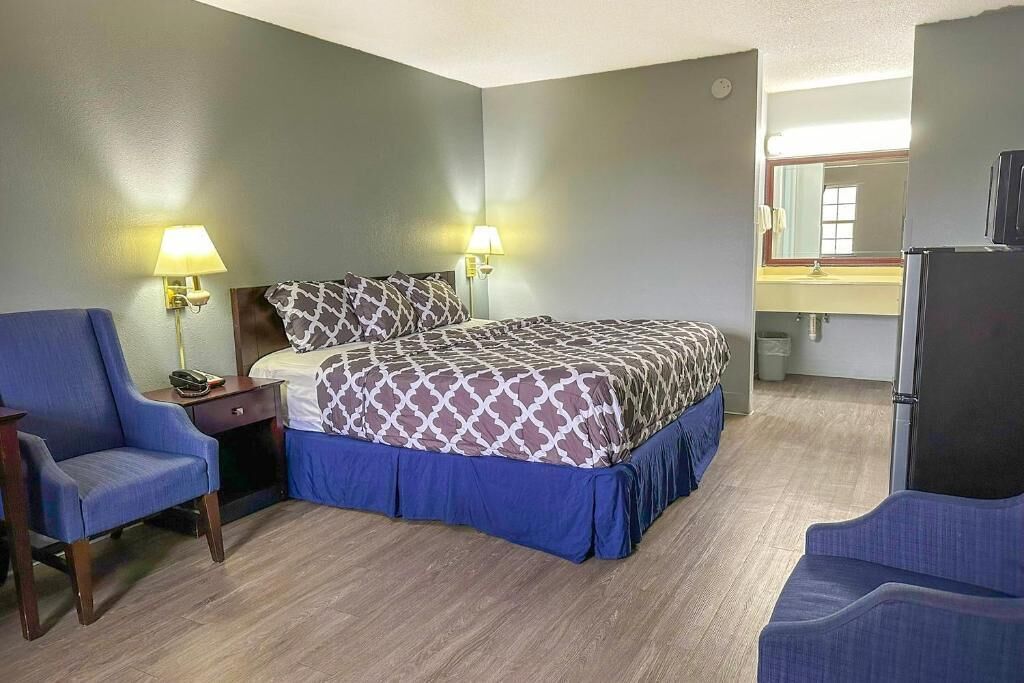 A hotel room with a bed, two bedside lamps, a desk with a chair, a refrigerator, and a mirror.