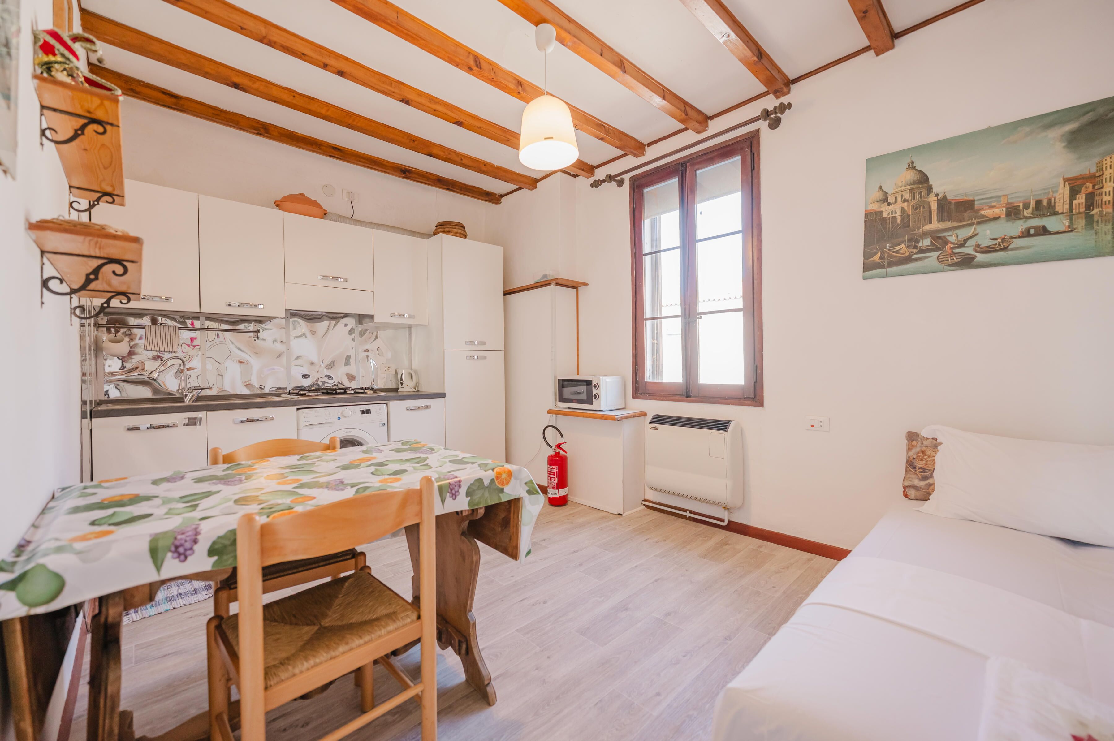 A kitchen with wooden beams, a dining table with a fruit pattern tablecloth, a bed, and a painting of a cityscape.