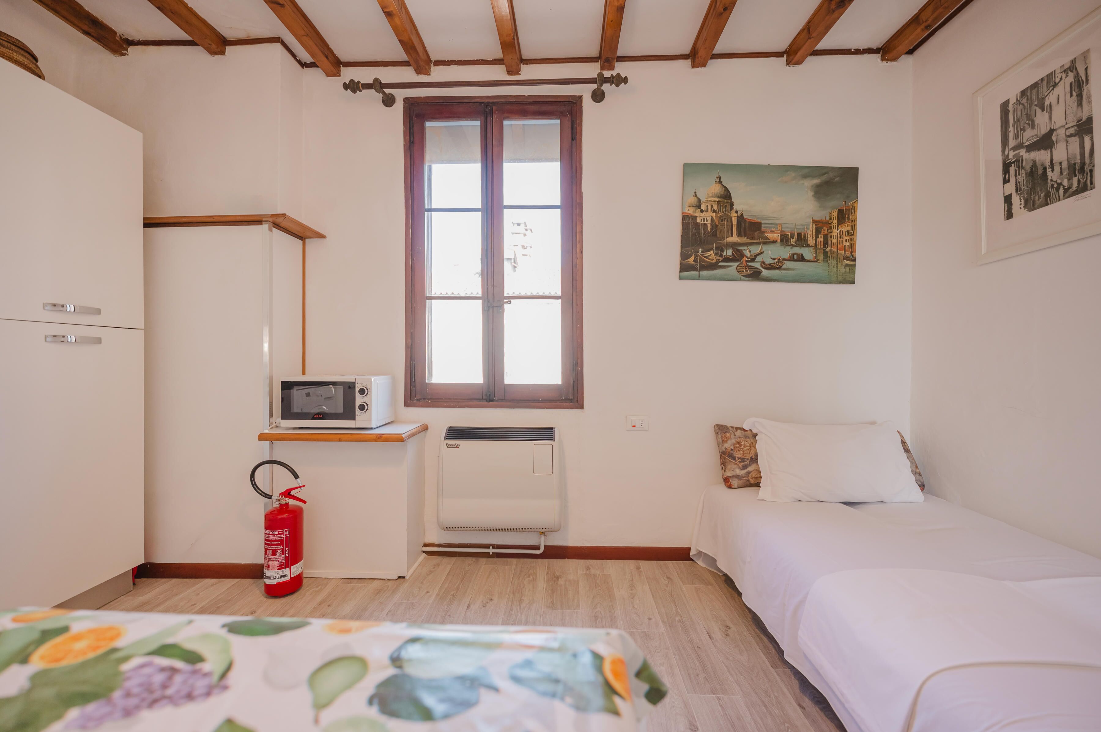 Una habitación con una cama, una mesita con microondas, un extintor y una ventana que da a vistas de un edificio.