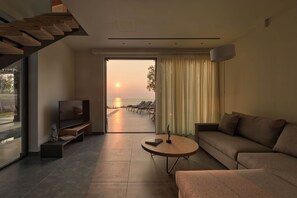 Ein modernes Wohnzimmer mit Fernseher, Sofa und Couchtisch, ein großes Fenster mit Blick auf den Sonnenuntergang und Strandstühle draußen.
