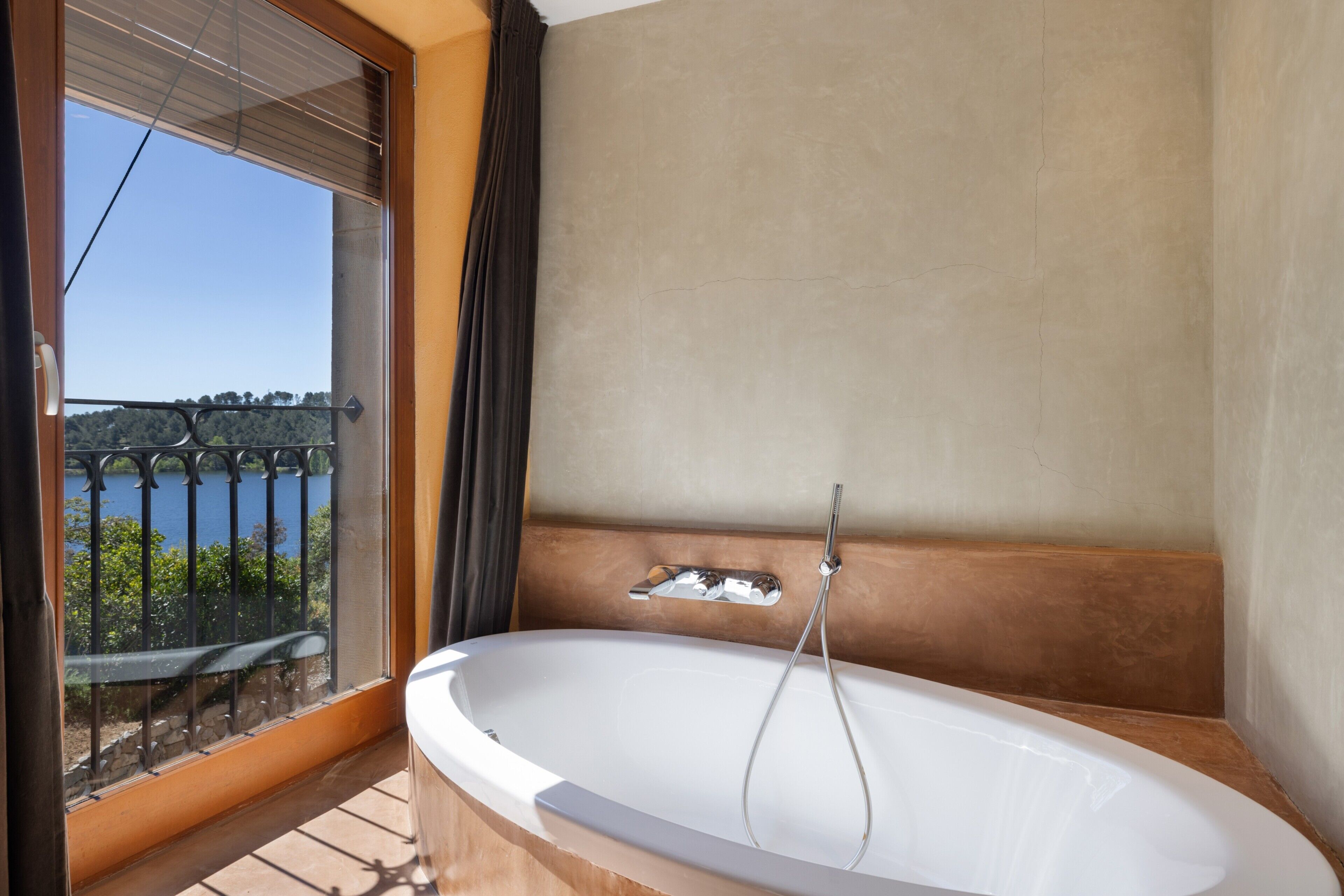 Un baño con bañera, una ventana que ofrece vistas a zonas verdes y una masa de agua, y un balcón con barandilla.