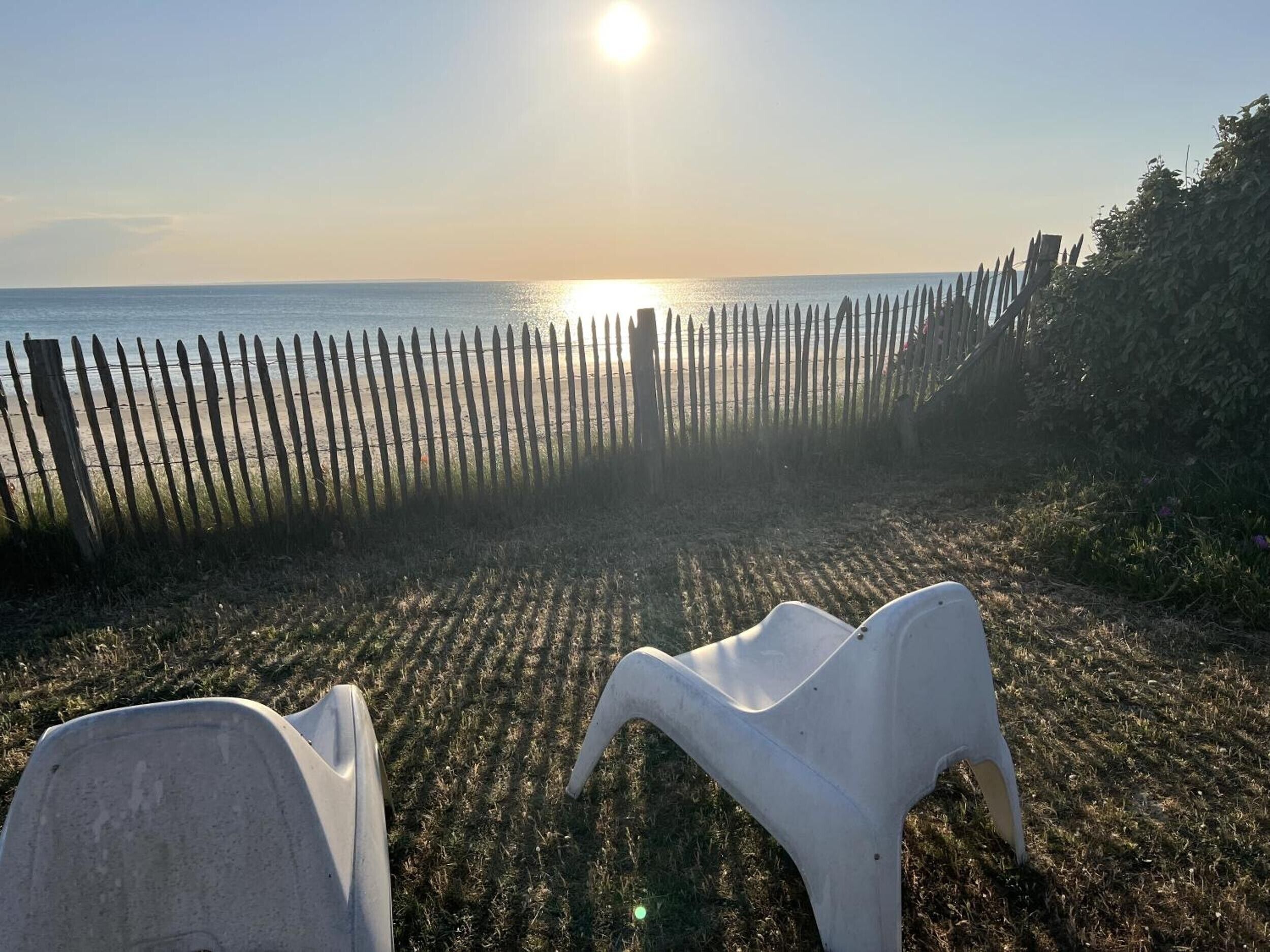 Blick auf einen Strand mit einer Holzlattenveranda, wolkenlosem Himmel und zwei weißen Stühlen.