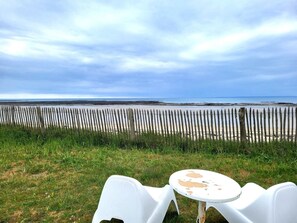 Uitzicht op een strand met een houten omheining, een gazon, en een witte plastic tafel en stoel.