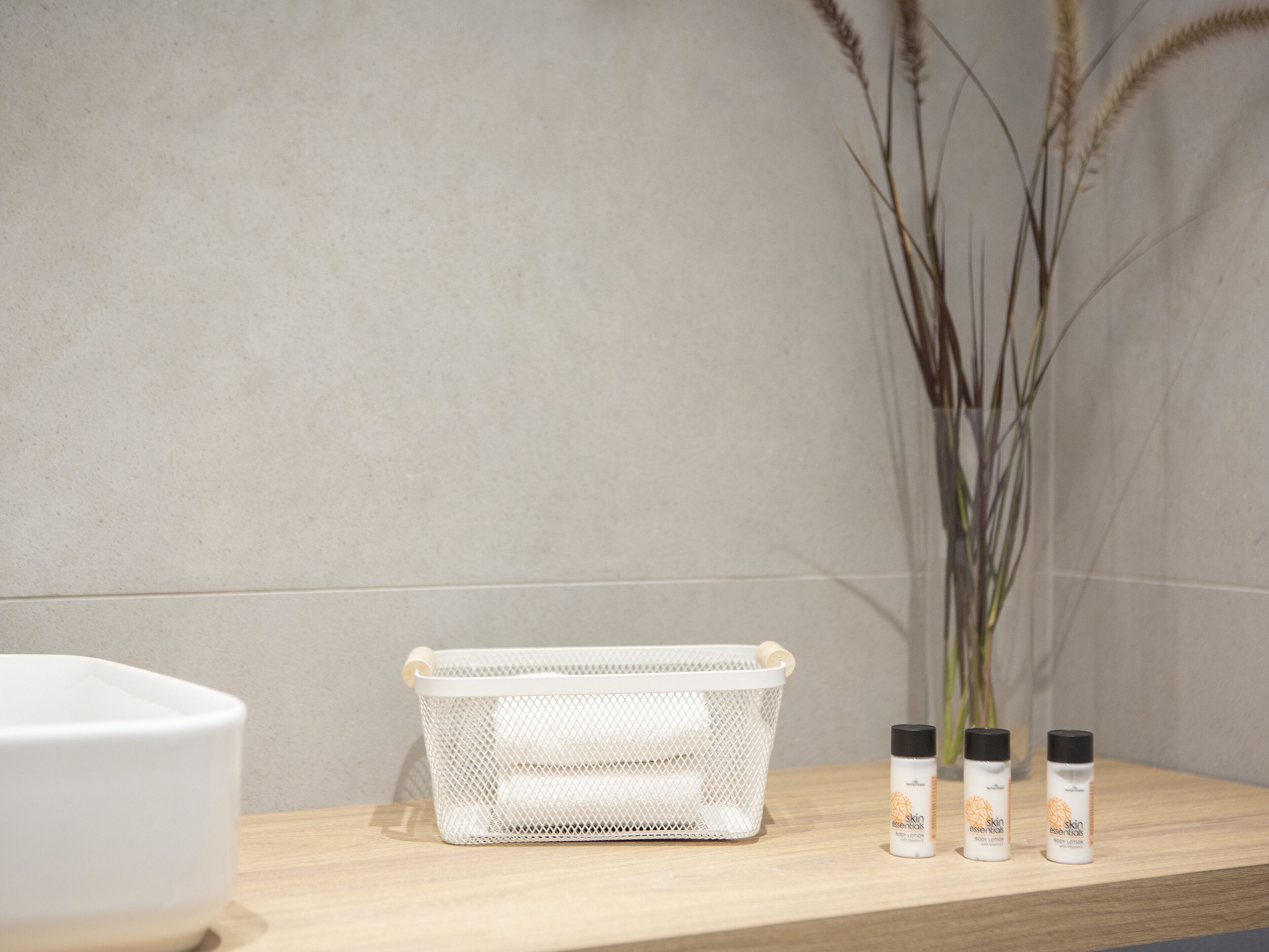 A bathroom with a wooden shelf, a white mesh basket, and two bottles of skin essentials body lotion.