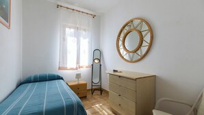 Een slaapkamer met een bed, een kaptafel, een stoel, een spiegel en een raam met dunne gordijnen.