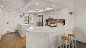 A modern kitchen with white cabinetry, a central island with stools, a microwave, and a stainless steel refrigerator. - Loggerhead Cay 362: Updated Coastal Charm – Steps to Beach with Gulf Views & New (Sanibel)