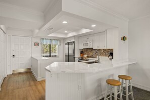 A modern kitchen with white cabinetry, a central island with stools, a microwave, and a stainless steel refrigerator. - Loggerhead Cay 362: Updated Coastal Charm – Steps to Beach with Gulf Views & New (Sanibel)