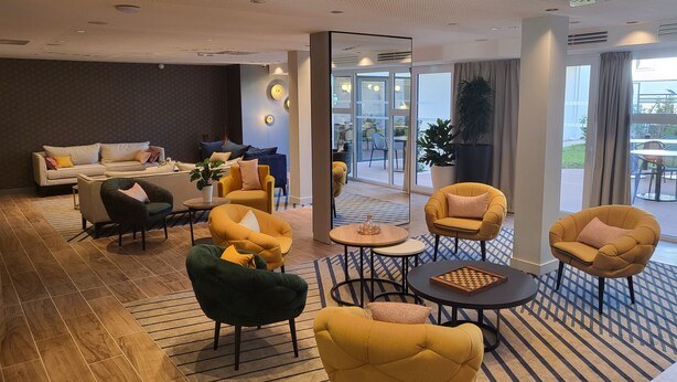 Una hall d'albergo moderna con un divano, poltrone, un tavolino da caffè e un grande specchio che riflette l'ambiente.
