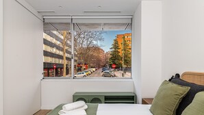 Un dormitorio con un ventanal grande, una cama, una mesita de noche y vista a la calle.