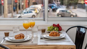 Una mesa de desayuno con croissants, fruta y bebidas, vista desde una ventana con vista a una calle con coches.