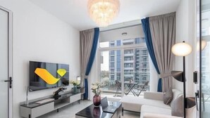 A modern living room with a flat-screen TV, a sofa, a coffee table, and a balcony with a view of buildings. - Vogue | Pearlz by Danube near Ibn Battuta Mall (Dubai)