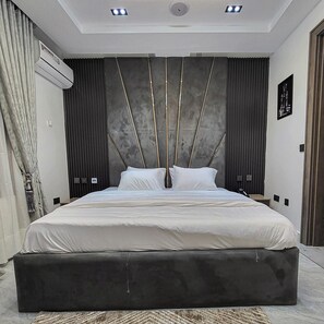 A modern bedroom with a large bed, dark curtains, an air conditioning unit, and a ceiling light fixture. - JBL HOTEL (Abuja)