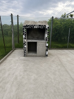 A concrete outdoor grill with a mosaic pattern, surrounded by a chain-link fence and greenery.