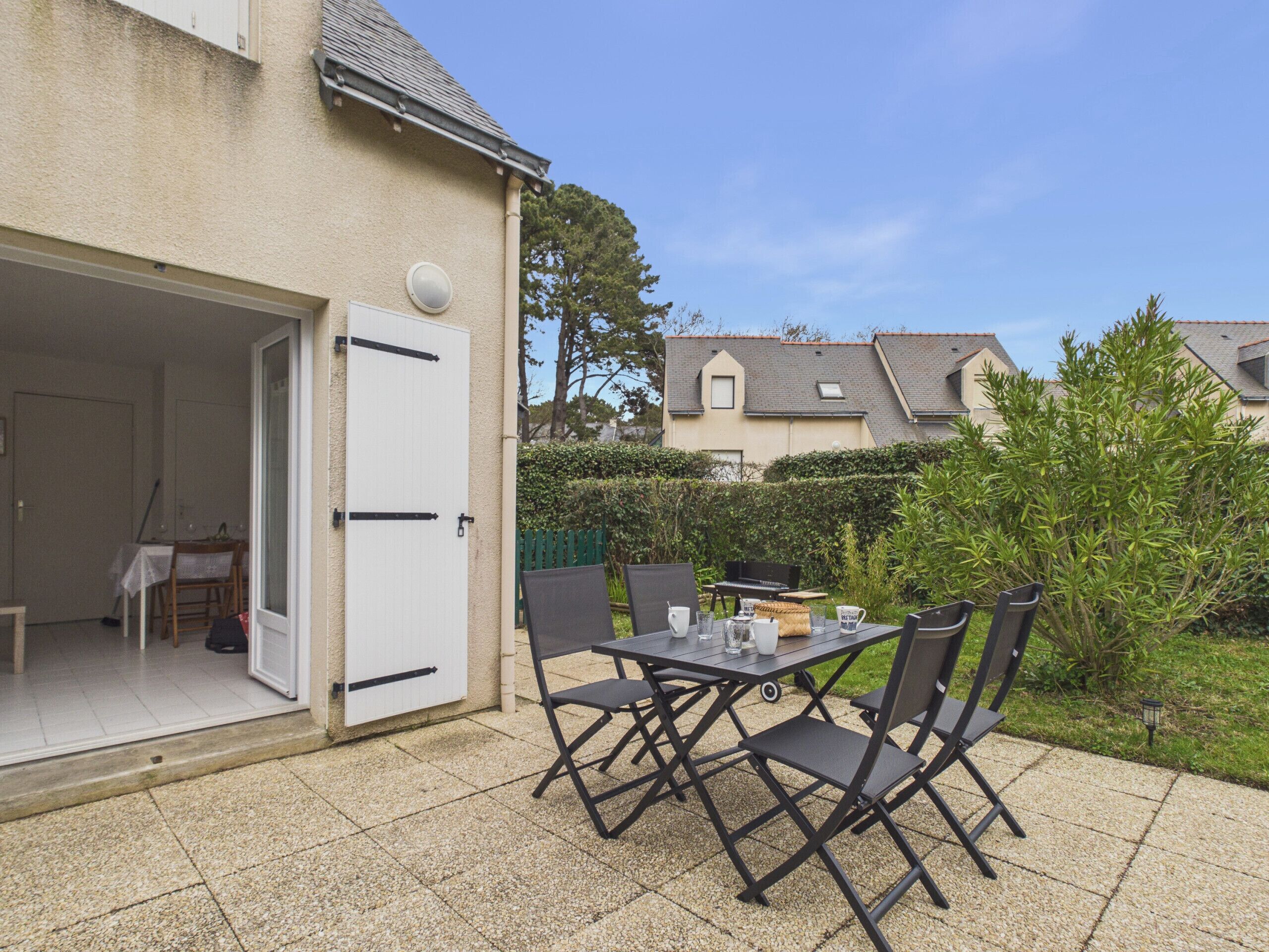 An outdoor patio with a table and chairs, a door leading to a room, and a hedge in the background.