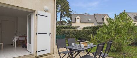 An outdoor patio with a table and chairs, a door leading to a room, and a hedge in the background.