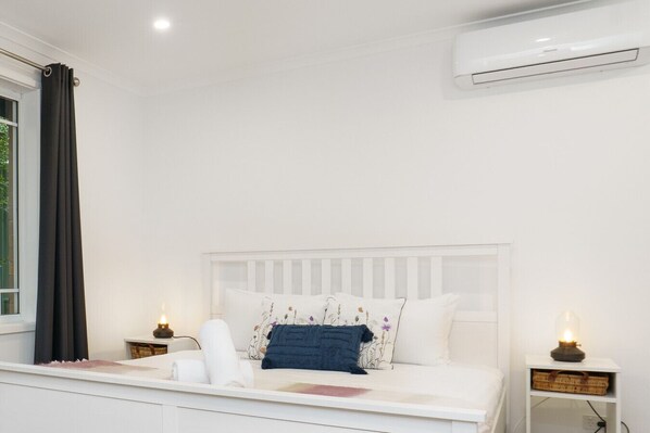 A white bed with a headboard, two bedside tables with lamps, and an air conditioning unit on the wall.